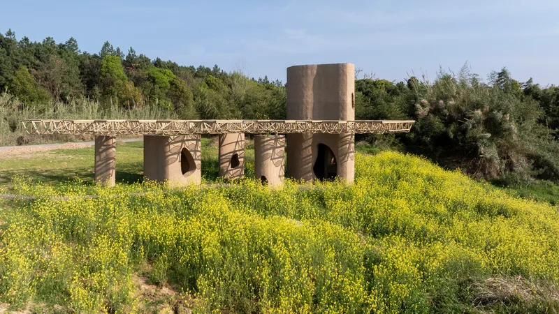 Cylindrical earthen columns with arched openings beneath a latticed canopy surrounded by blooming yellow wildflowers