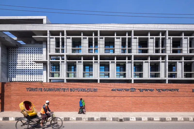 Street view of the perforated white facade with horizontal concrete frame above red brick boundary wall