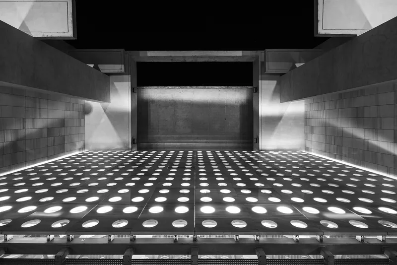 Upward view of perforated metal floor grate with circular openings and backlit concrete wall panel