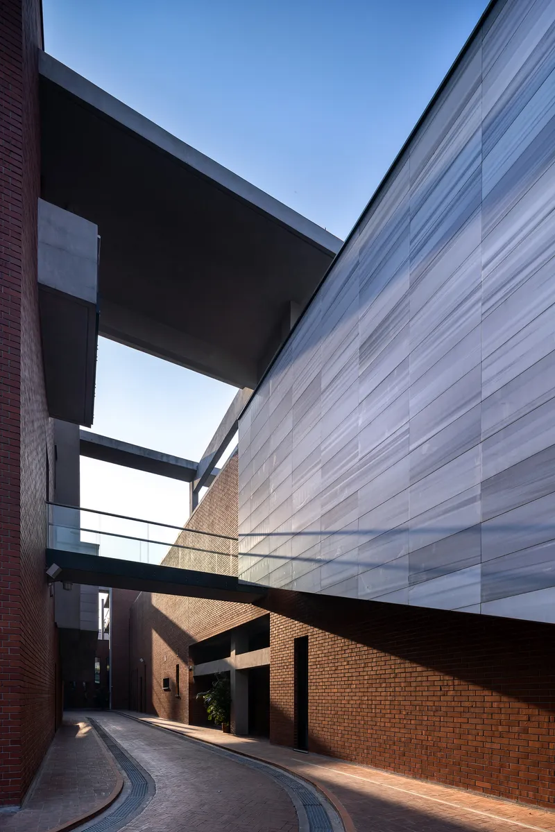 Curving paved lane between brick facades and a translucent panel wall connected by concrete bridges
