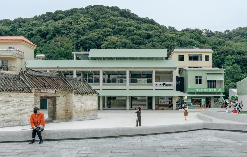 Paved plaza with a traditional tile-roofed building beside a pale green multi-storey structure backed by forested hills