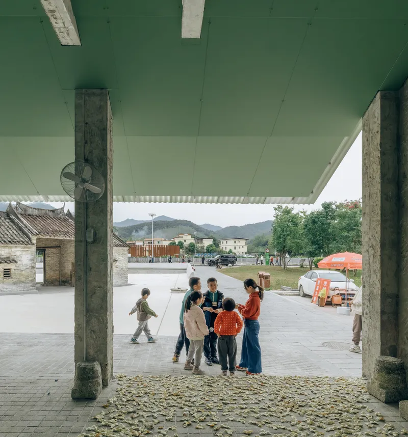Covered terrace with exposed concrete columns and mint green soffit overlooking children gathered on cobblestones
