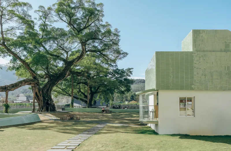 White plastered pavilion with green ceramic tile roof beside a stepping-stone path and old fig tree