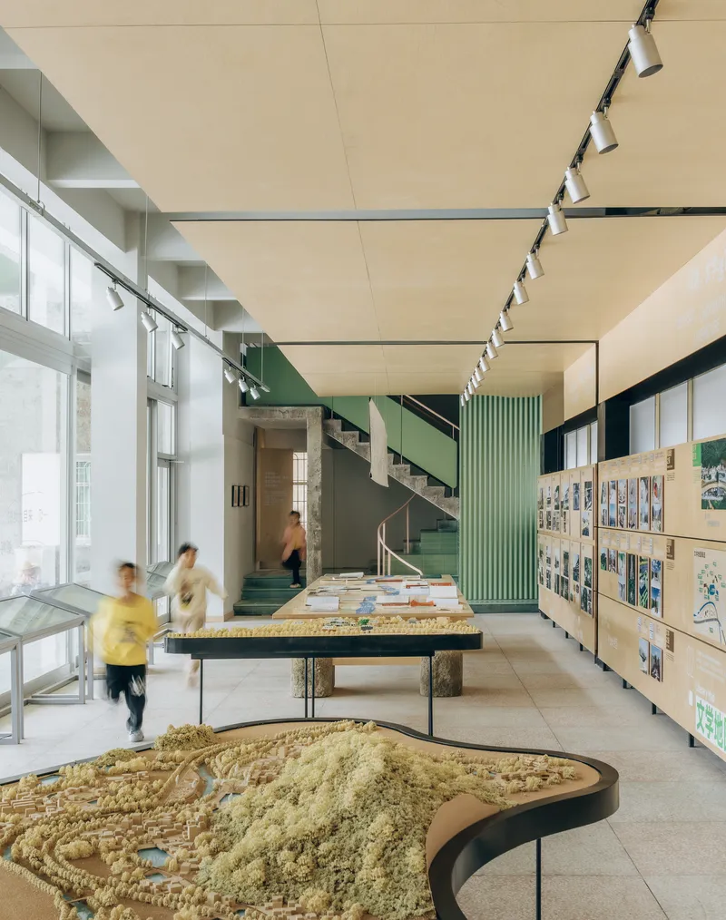 Exhibition hall interior with topographic models on tables, plywood display walls, and a green-sided staircase beyond