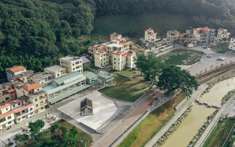 Aerial view of the village center with the elongated green-roofed building and concrete plaza beside the river