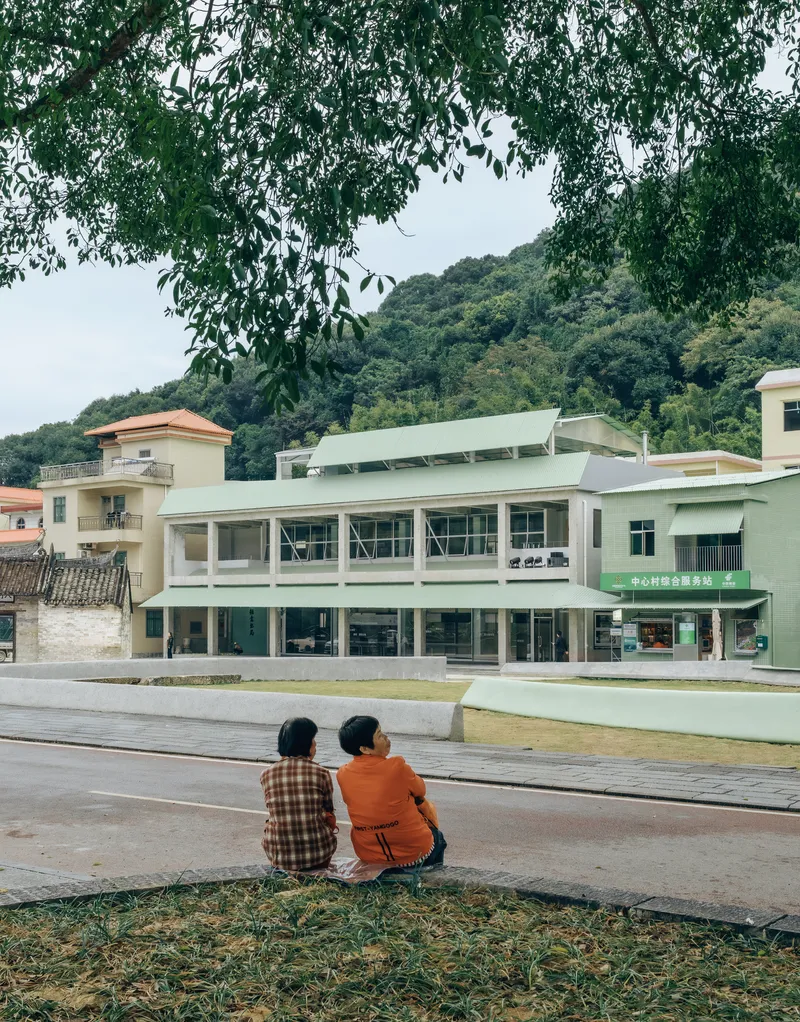 Two figures sitting on the stepped concrete edge watching the green-roofed building across the lawn
