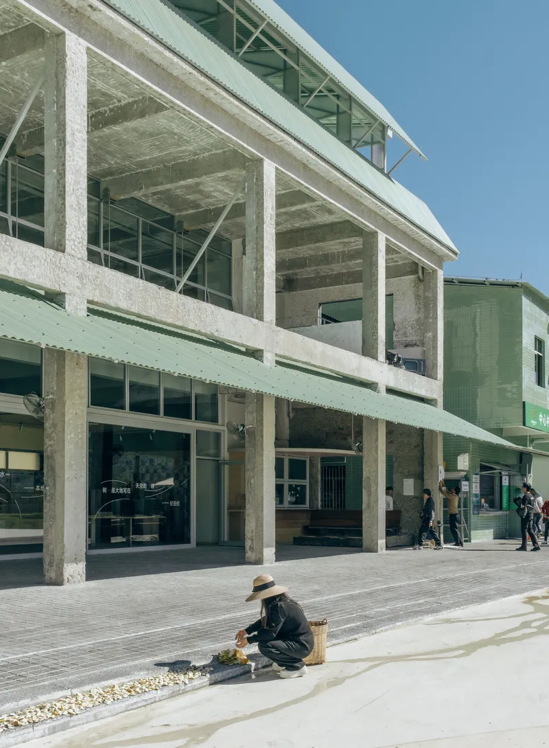 Open ground floor with exposed concrete structure and figure crouching by a gravel bed in sunlight