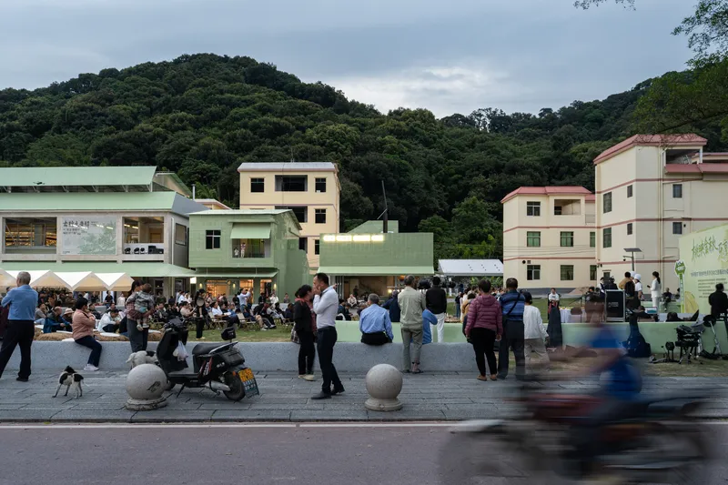 Plaza view of the pale green pavilion with surrounding residential buildings and a forested hillside under cloudy skies