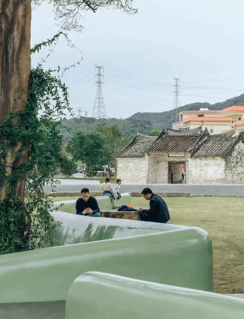 Curved pale green seating elements with visitors amid trees and a historic stone building beyond
