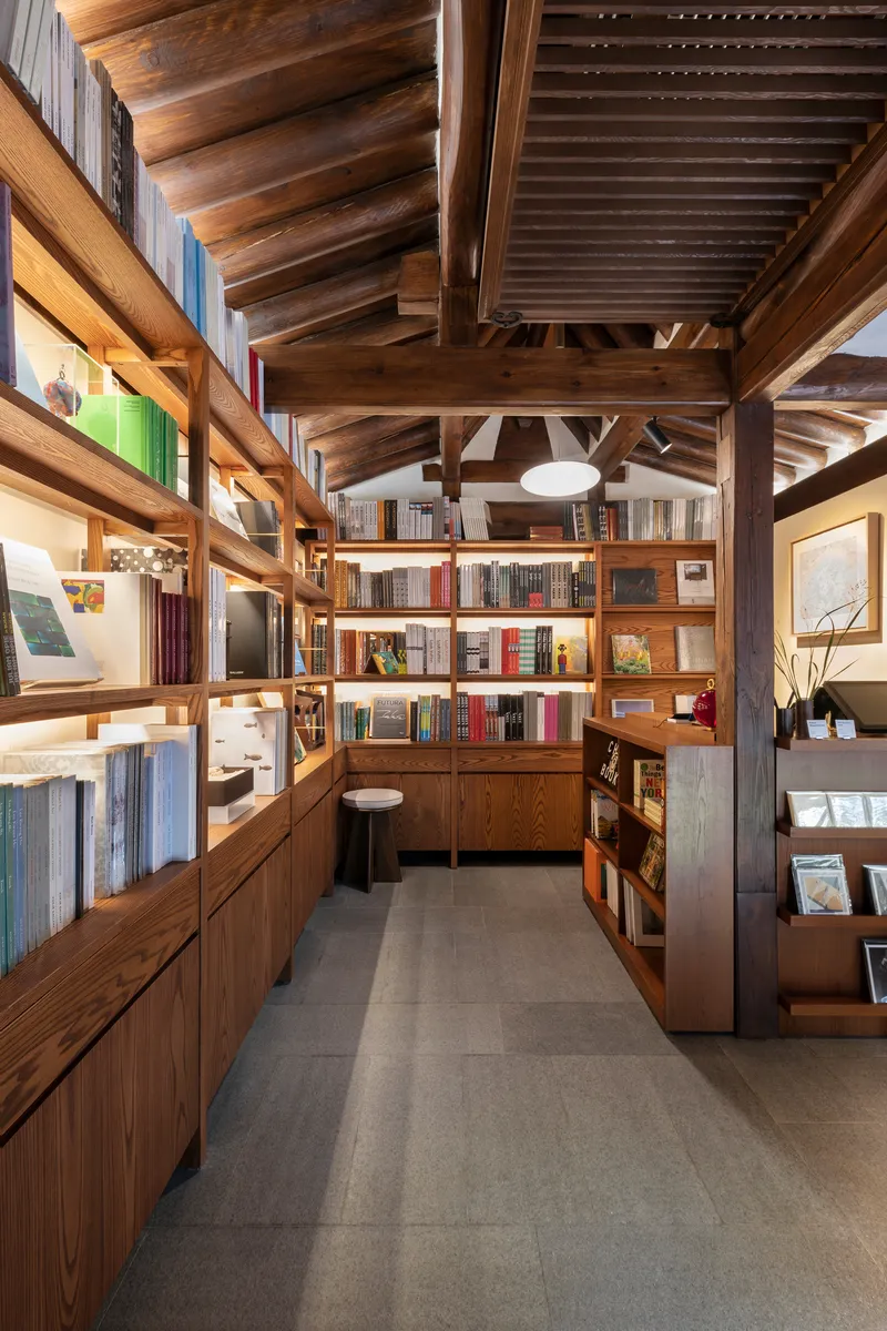 Library corridor with illuminated wooden bookshelves beneath heavy timber rafters and steel tension rods