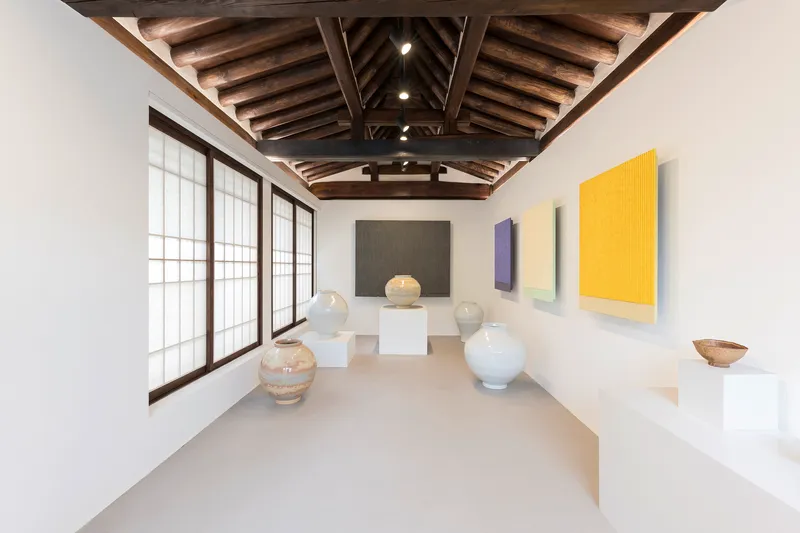 Gallery room with white ceramic vessels on pedestals and colorful paintings beneath timber truss ceiling
