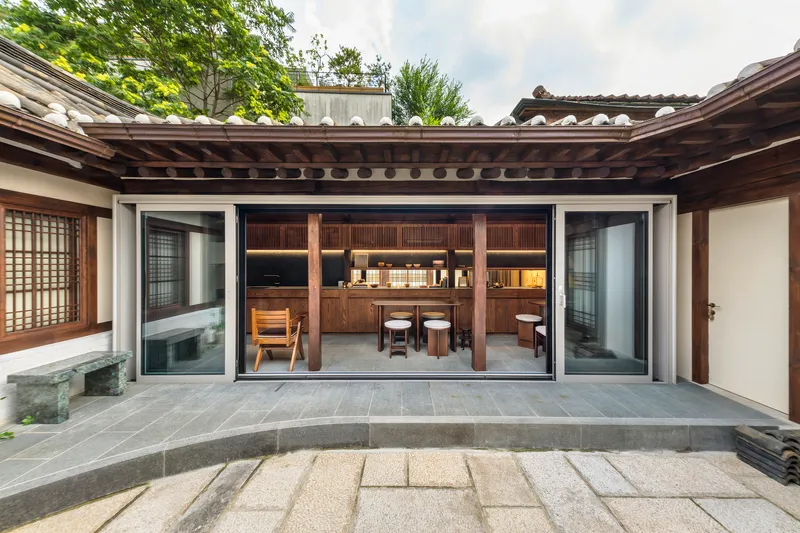 Courtyard view through open sliding glass doors into a timber kitchen with exposed rafter ceiling