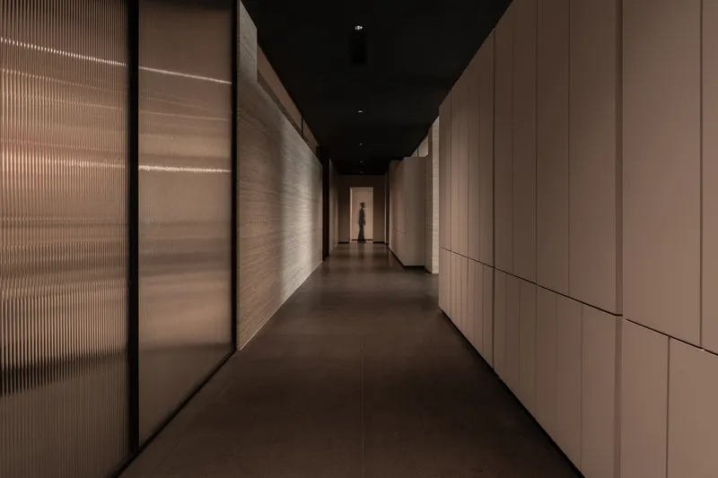 Narrow hallway with ribbed translucent glass walls and wood paneling leading to a distant silhouette