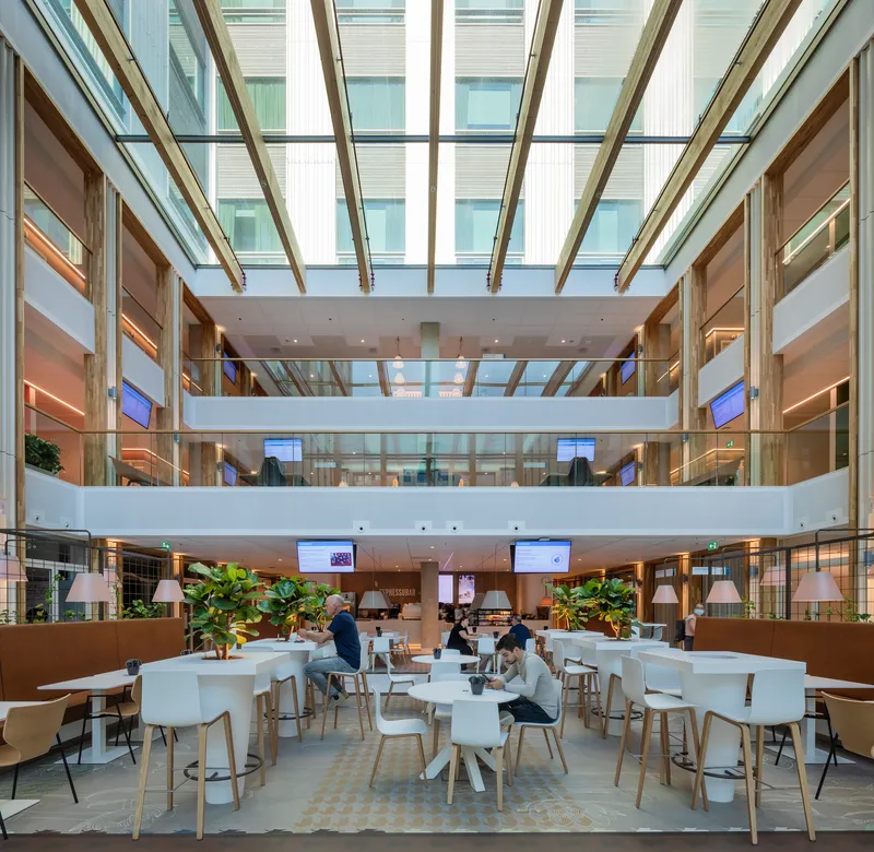 Multi-level atrium cafe with angled glass roof and people seated at white tables