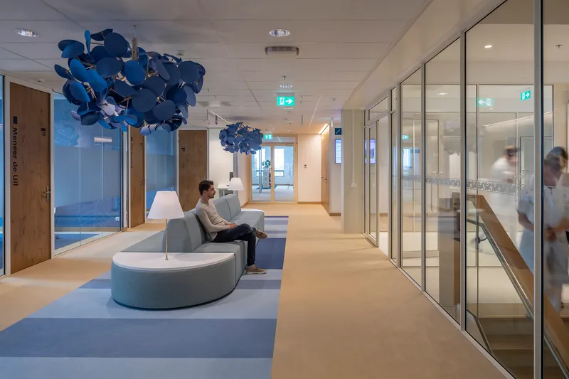 Corridor with curved seating, blue sculptural pendant lights, and glass-walled offices with a person working