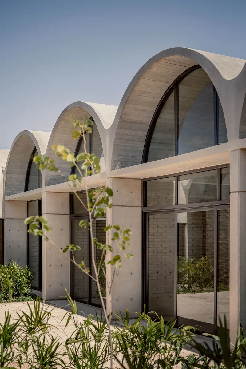 Facade with repeating vaulted concrete shells above arched glazing and young saplings in foreground planting