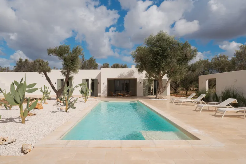 Courtyard pool flanked by white stucco volumes with olive trees and cacti under a partly cloudy sky