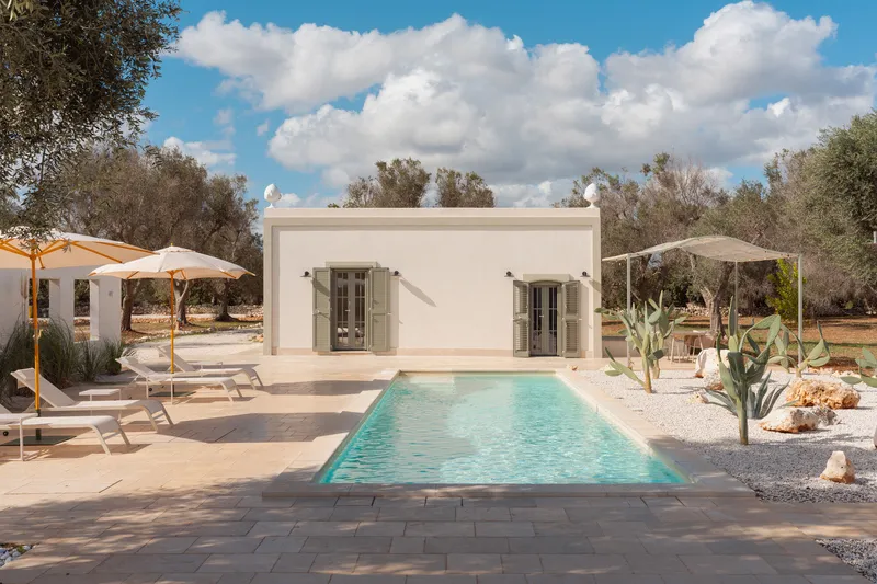 Pool terrace with stone pavers framed by white umbrellas and scattered cacti beneath olive trees
