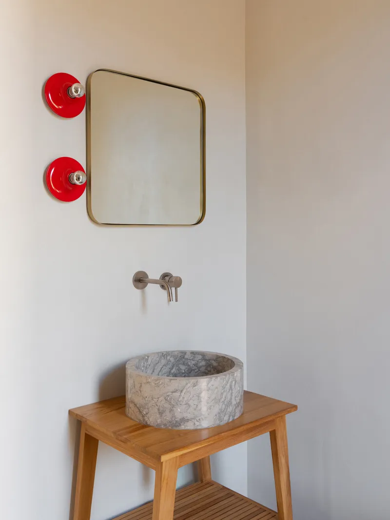 Circular stone basin on timber stool beneath brass-framed mirror and red ceramic wall sconces