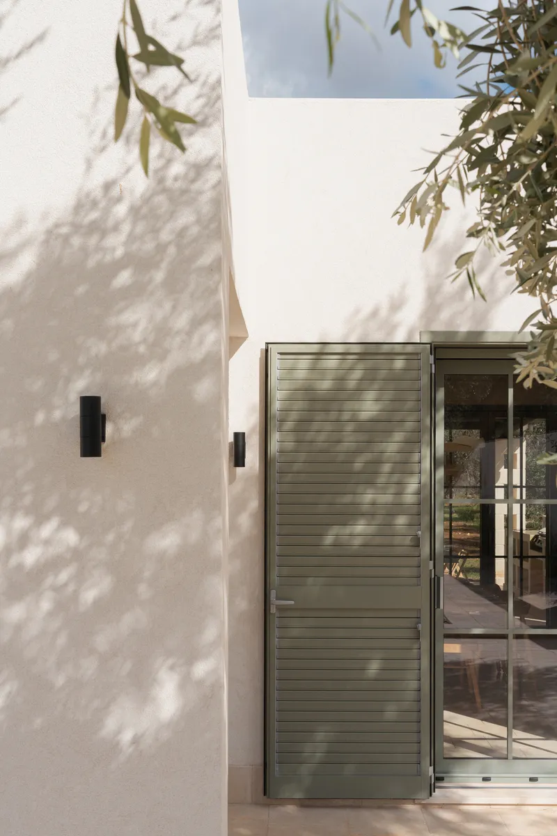 White facade with louvered green door and olive tree shadows cast across the wall