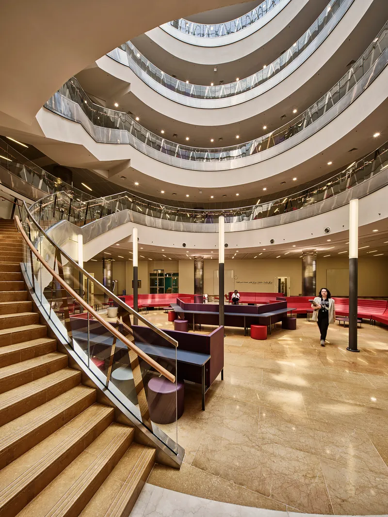 Multi-story atrium with curved balconies, timber and glass railings, and a person crossing the ground floor
