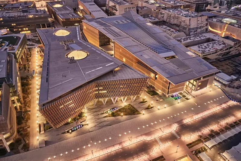 Aerial view at dusk of the complex showing illuminated plaza, lattice facade, and surrounding parking structures