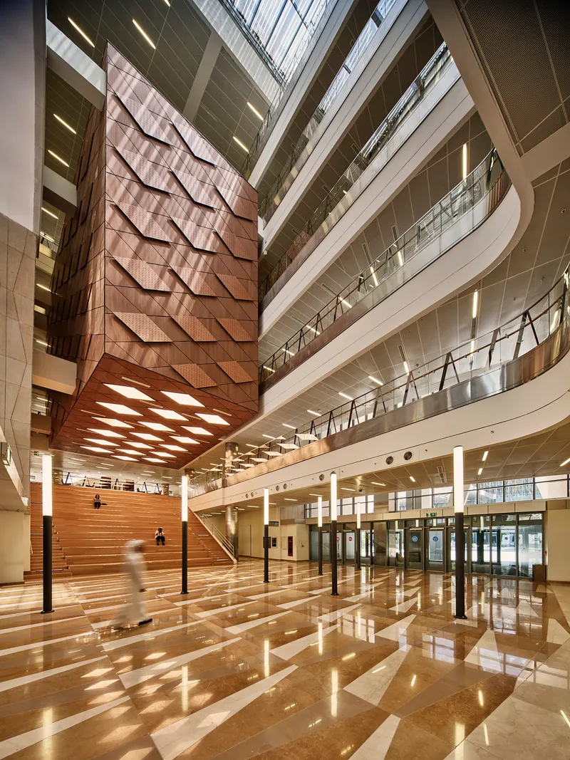 Multi-story atrium with timber-clad seating steps and polished stone floor reflecting skylight patterns