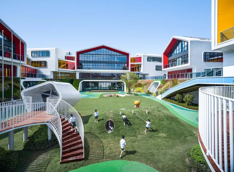 Elevated courtyard with grassed slopes and tubular slides where children play beneath white volumes with colored fenestration