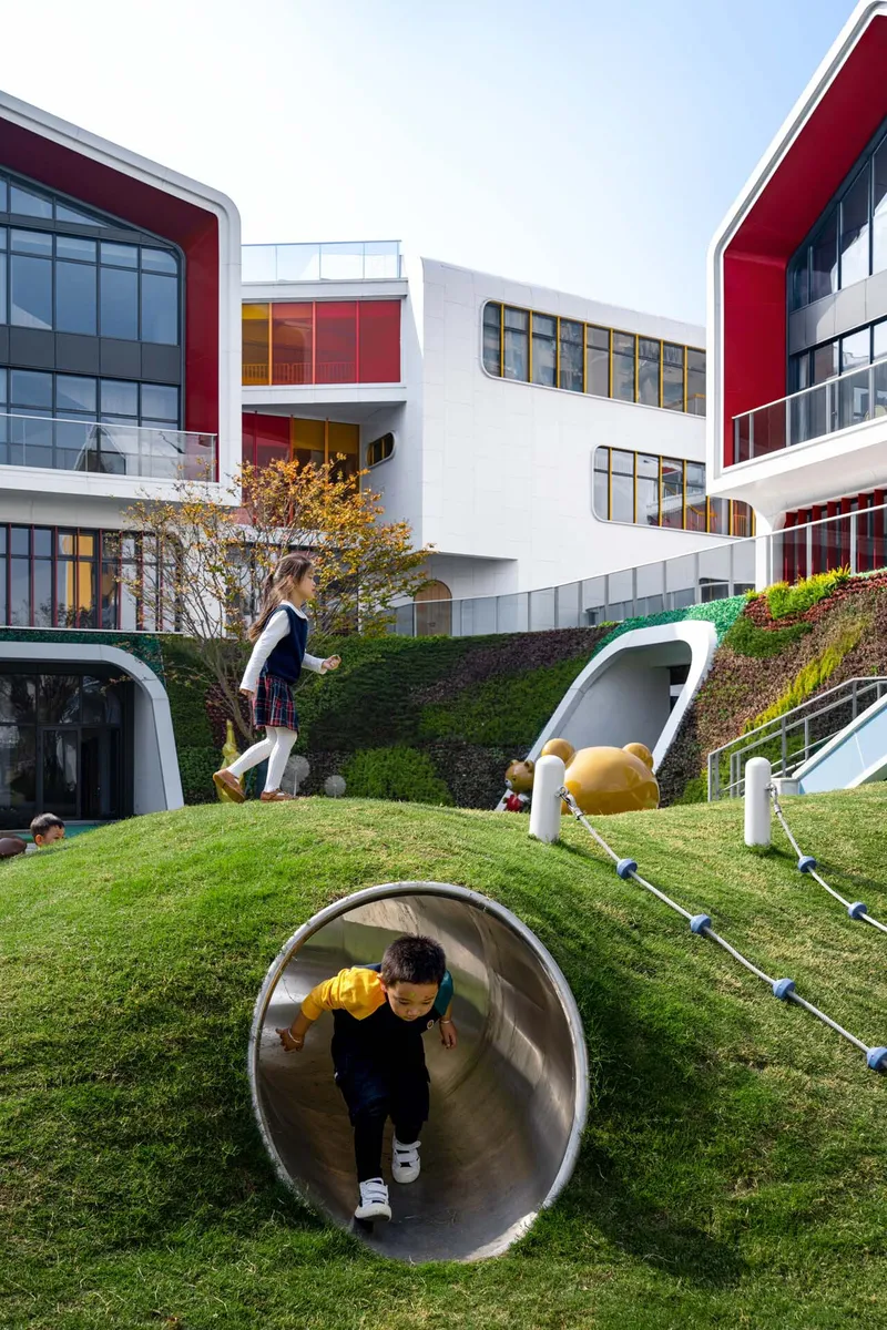 Child emerging from a white tunnel slide embedded in the grassed slope with students walking above