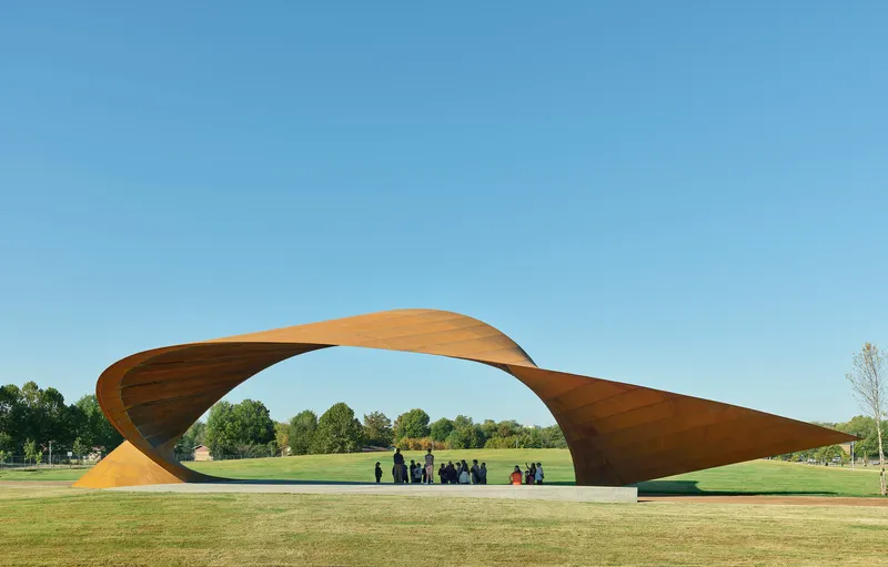 Twisted timber canopy spanning a lawn with visitors gathered beneath under clear blue sky