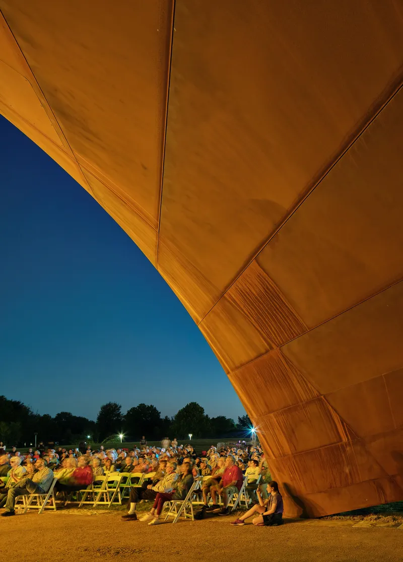 Evening performance with audience seated beneath the illuminated timber roof at dusk