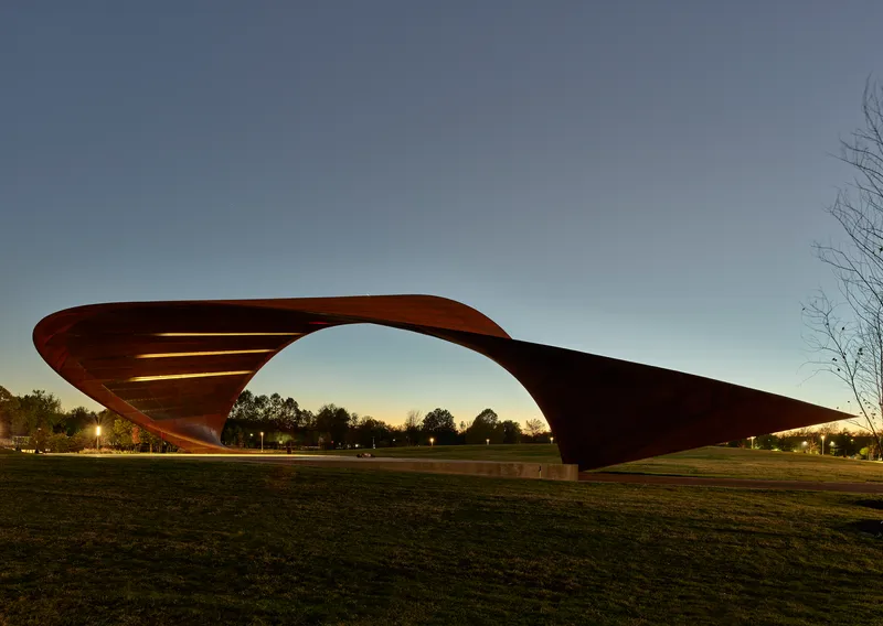 Sculptural canopy structure with sweeping curved roof silhouetted against the evening sky in a park