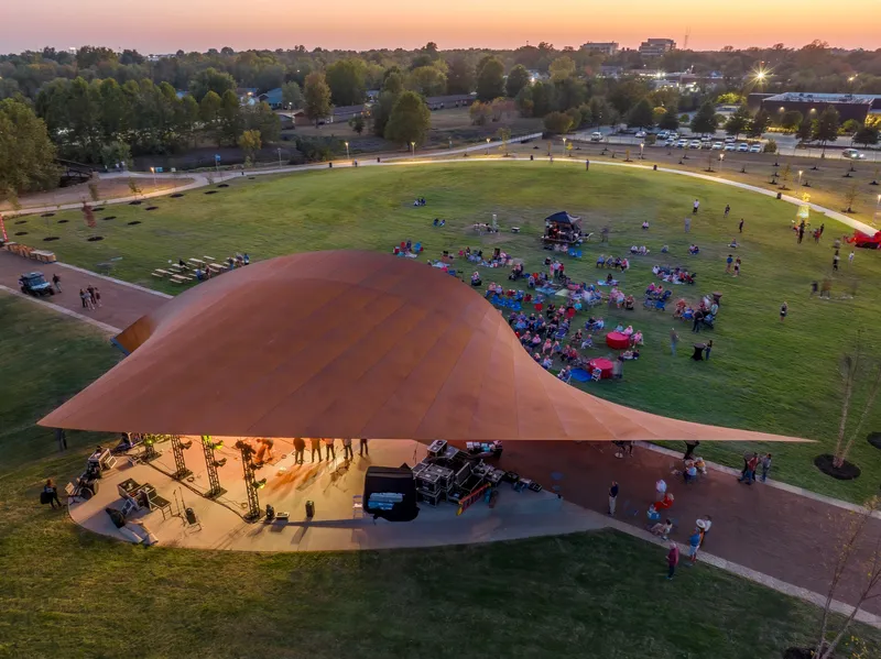 Aerial view of open-air pavilion with red metal roof sheltering a concert audience on the lawn at dusk