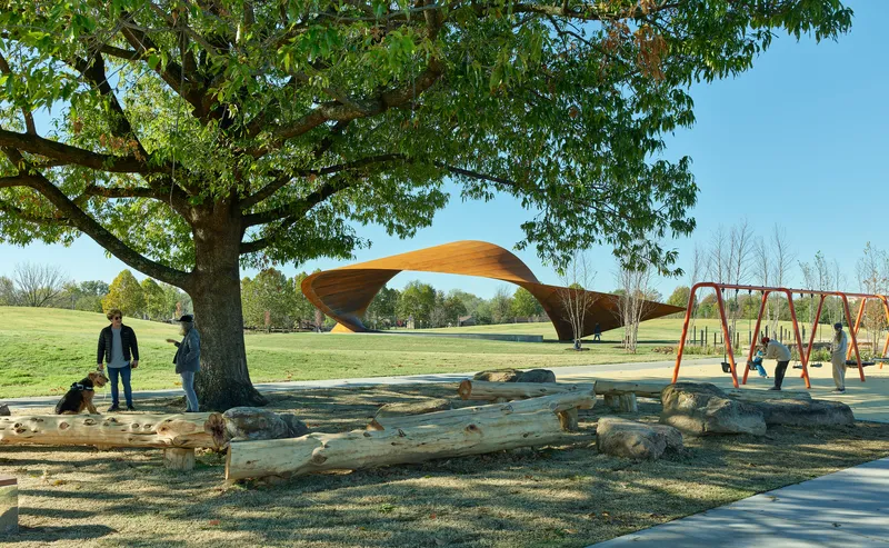 View across natural log play area toward the canopy structure framed by a mature tree