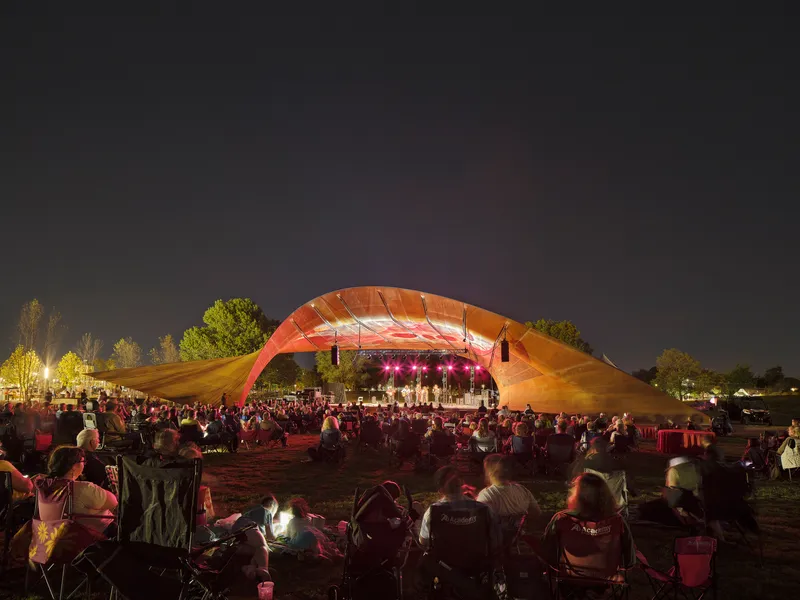 Nighttime concert beneath the illuminated shell roof with crowd gathered on the grass facing the stage