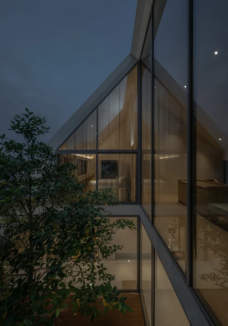 Exterior view of the angled glass facade with timber screening and planted trees at night under a rising moon