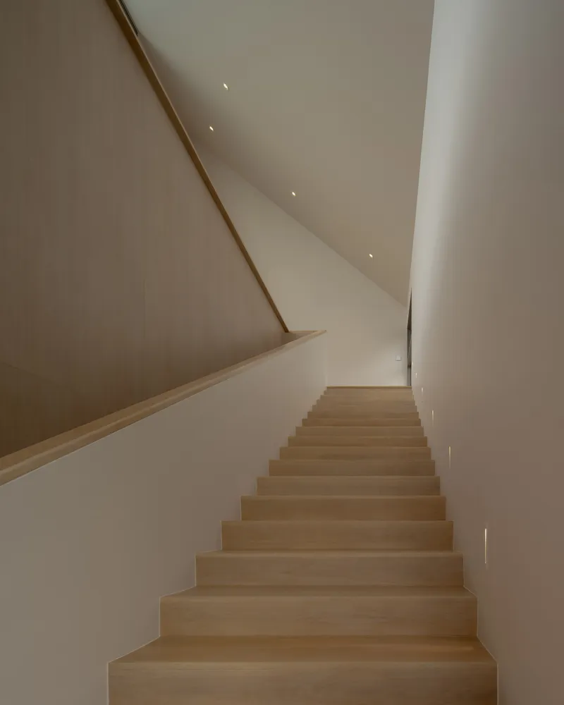 Light wood staircase ascending beneath a sloped white ceiling with recessed spotlights