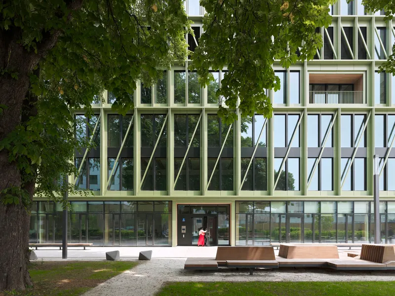 Ground-level entrance with green structural bracing behind mature trees and a timber bench in the plaza