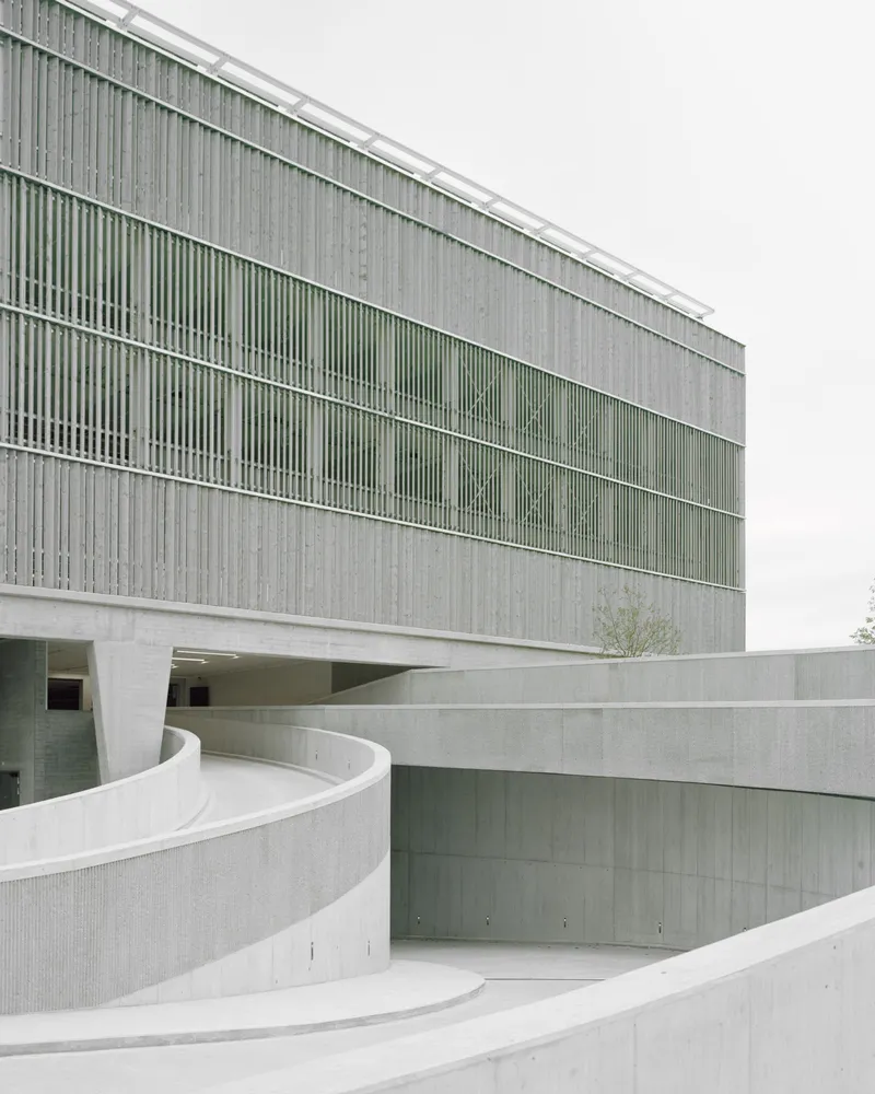 Curved concrete ramp walls beneath the louvered facade with a small tree emerging from the courtyard