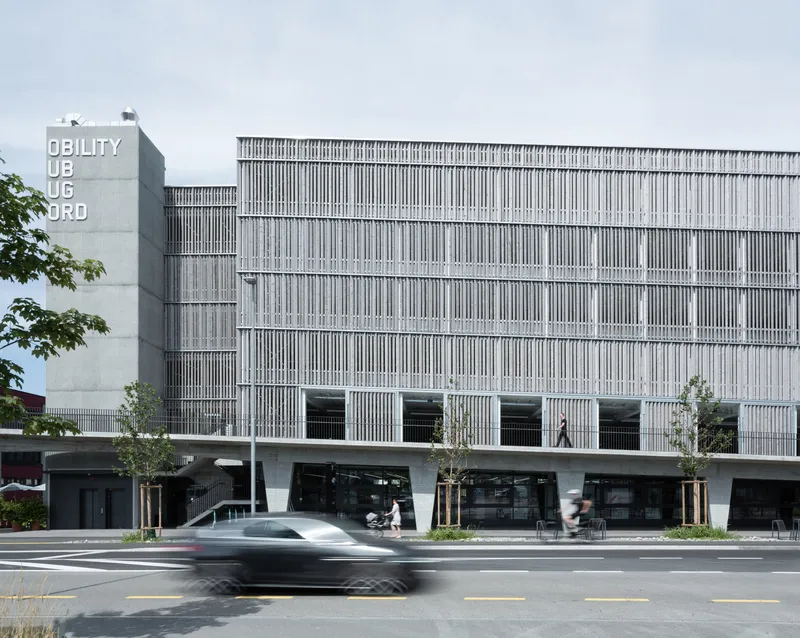 Facade with vertical louvers and canted concrete columns at street level as a car passes in the foreground