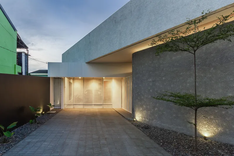 Entry courtyard with paver walkway leading to glazed doors beneath an illuminated concrete canopy at dusk