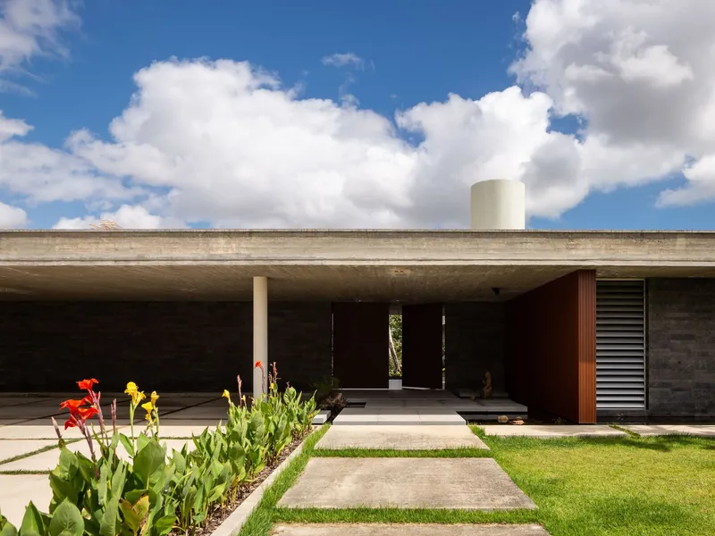 Concrete cantilevered roof sheltering an entrance portico with flowering canna lilies and grass lawn under white clouds