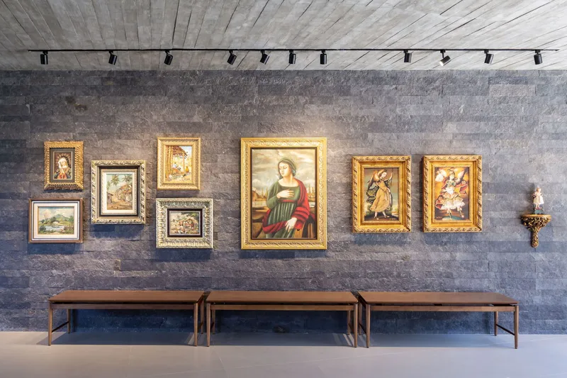Dark stone wall with collection of gilt-framed paintings above three low wooden benches and track lighting
