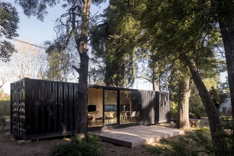 Dark corrugated metal volumes and glazed pavilion on a timber deck nestled among mature pine trees