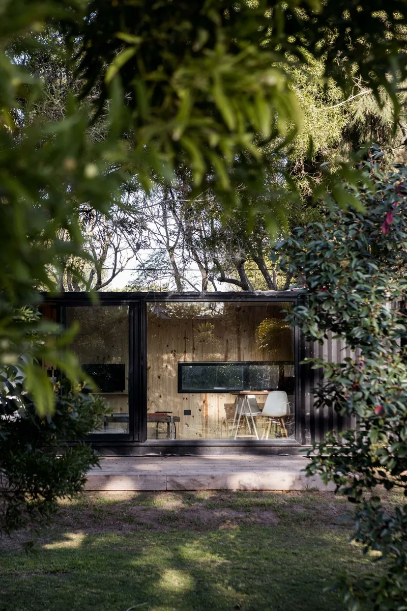 Glass-fronted pavilion framed by overhanging tree branches and surrounding garden foliage at dusk
