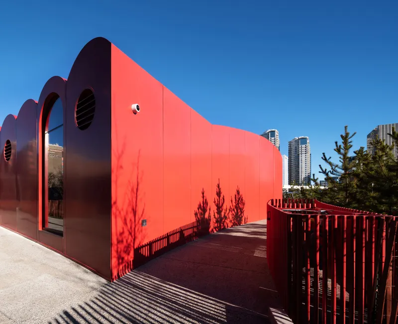 Red metal wall with circular window openings and tree shadows cast across the terrace