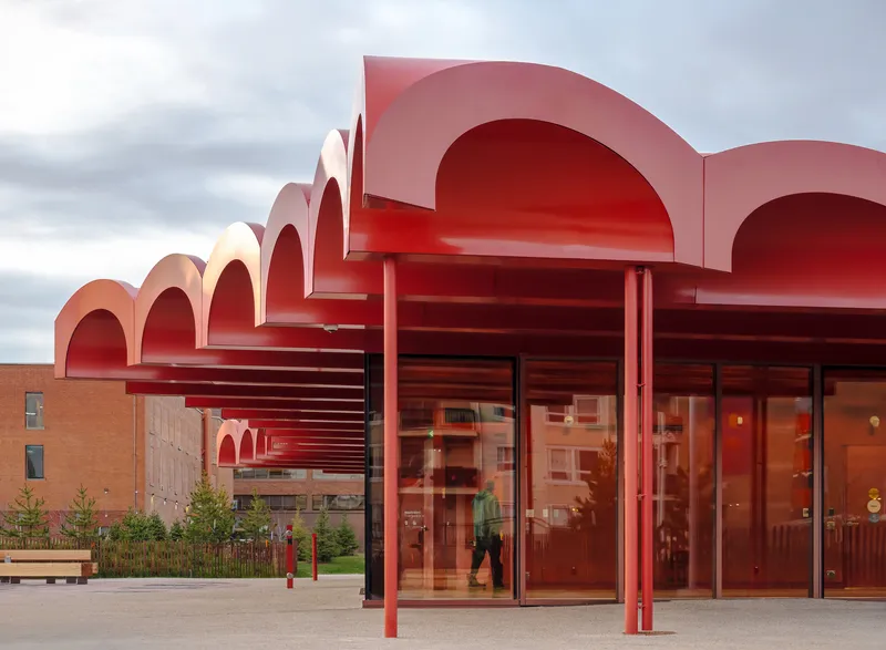 Red arched canopy with horizontal louvers above glass entrance doors beneath an overcast sky