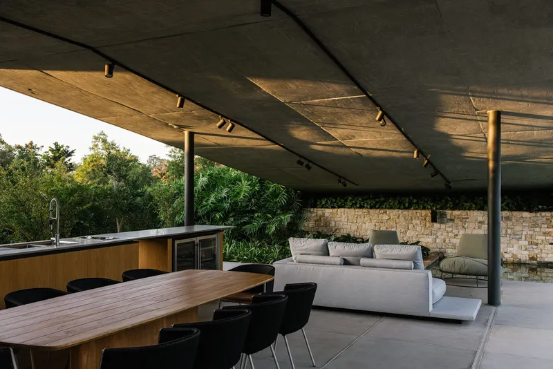 Covered terrace with concrete ceiling, steel columns, dining table and stone accent wall at sunset