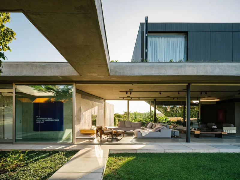 Open-air living terrace beneath exposed concrete soffits with lawn and pool deck in afternoon light