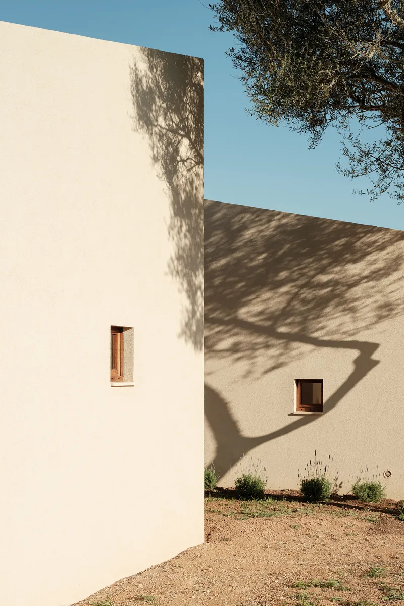 Corner junction of two white stucco walls with small timber-framed windows under tree shadow